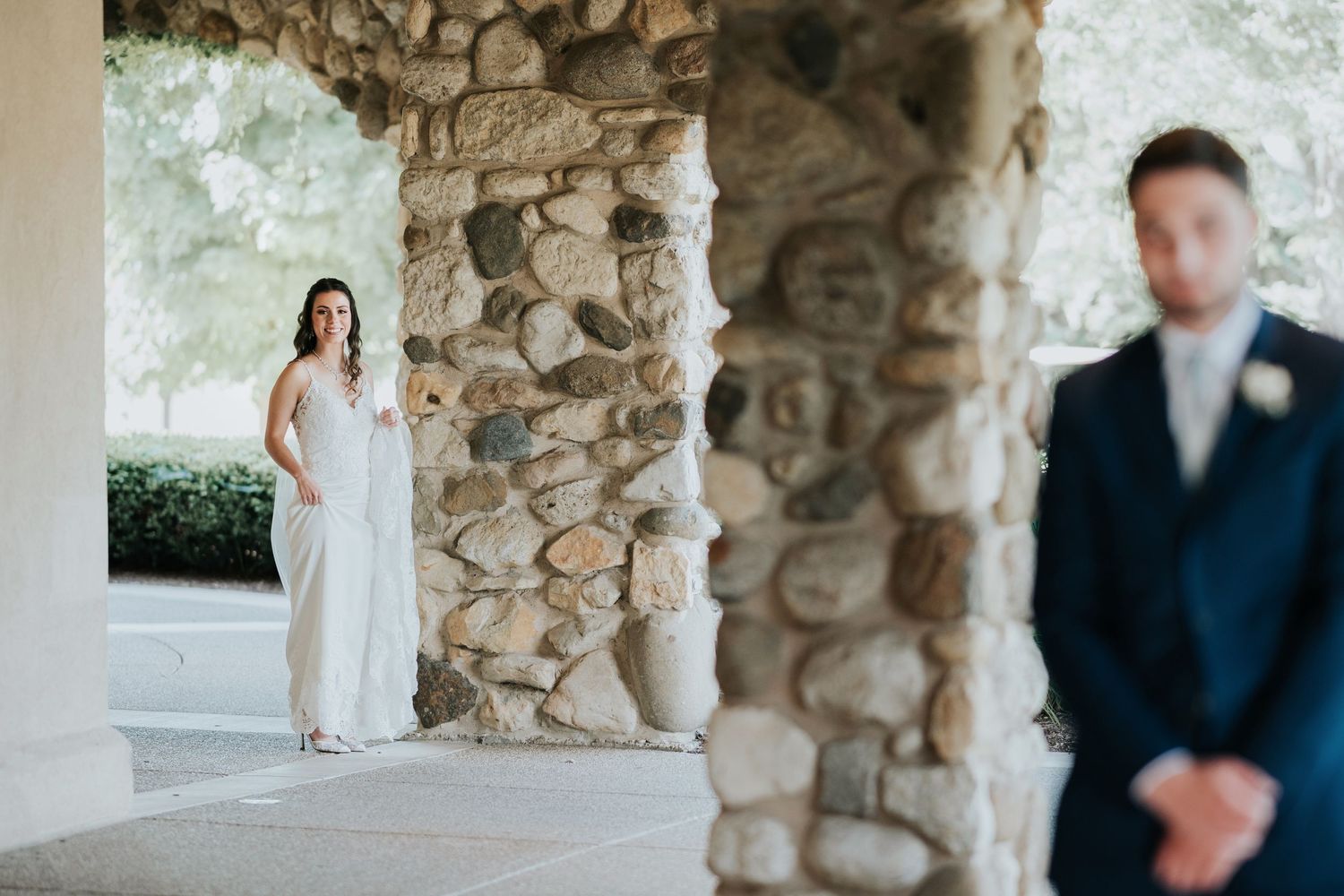 Bride in wedding dress walking under cobblestone arch for first look with groom stading in foreground.