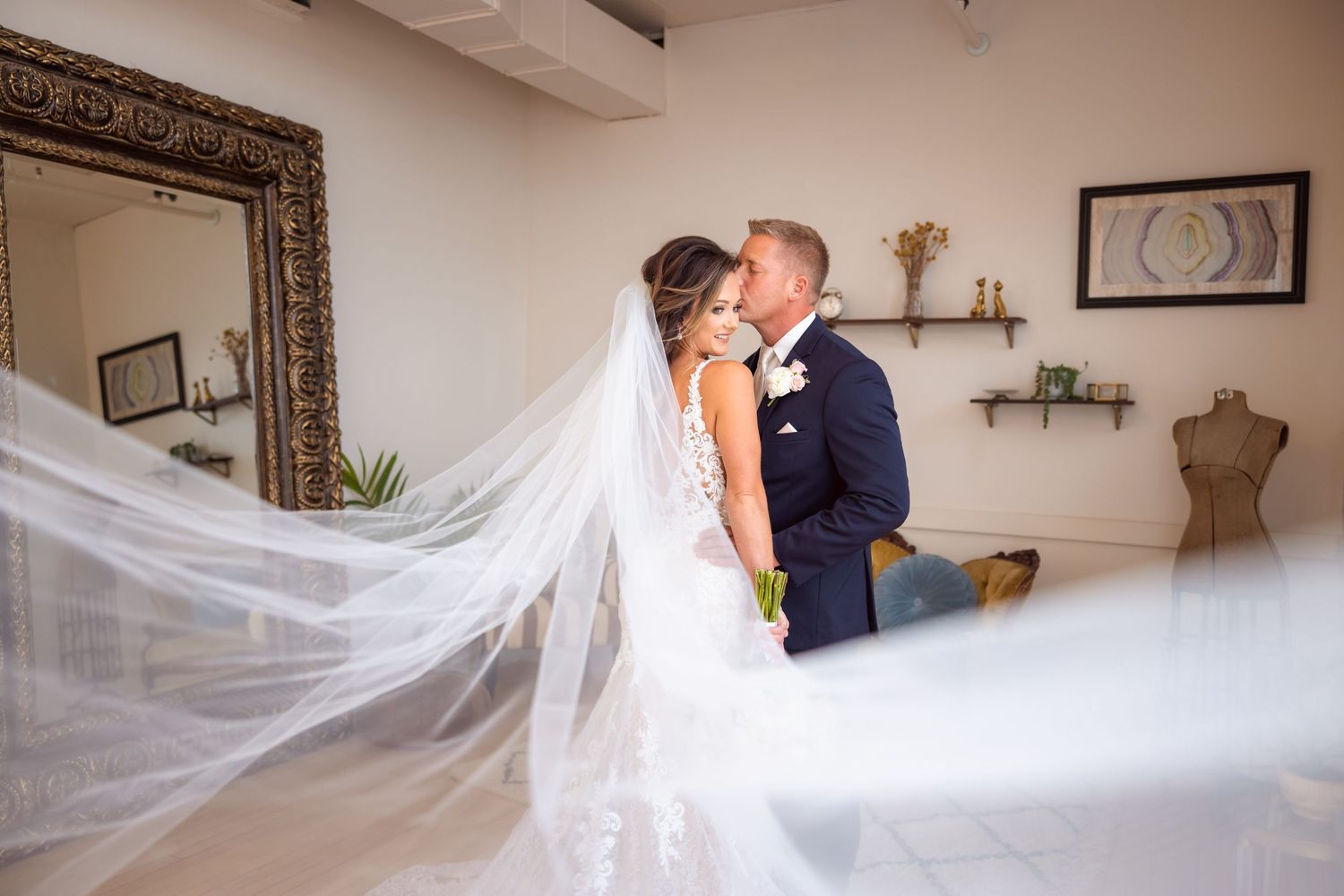 Wedding couple embraces in elegant indoor setting with flowing white veil and ornate mirror.