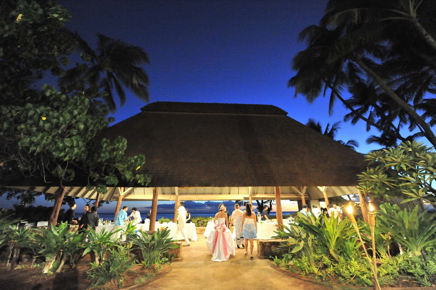 Elegant open-air thatched pavilion illuminated at night with wedding guests gathered along a tropical pathway.