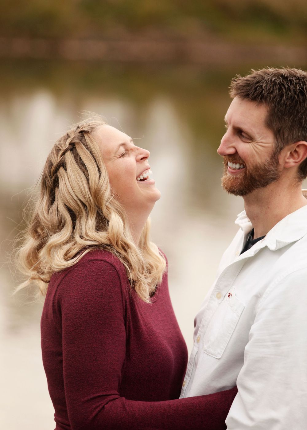 Happy couple in burgundy and white share joyful moments during lakeside photoshoot.