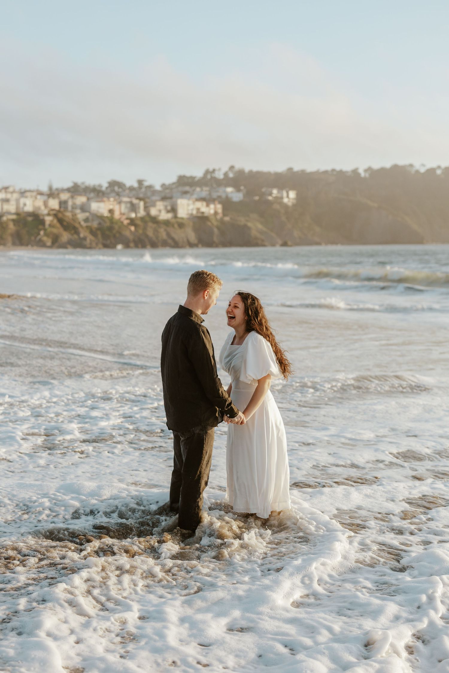 an engaged couple laughing and standing in the ocean at Baker Beach in San Francisco during their engagement photoshoot