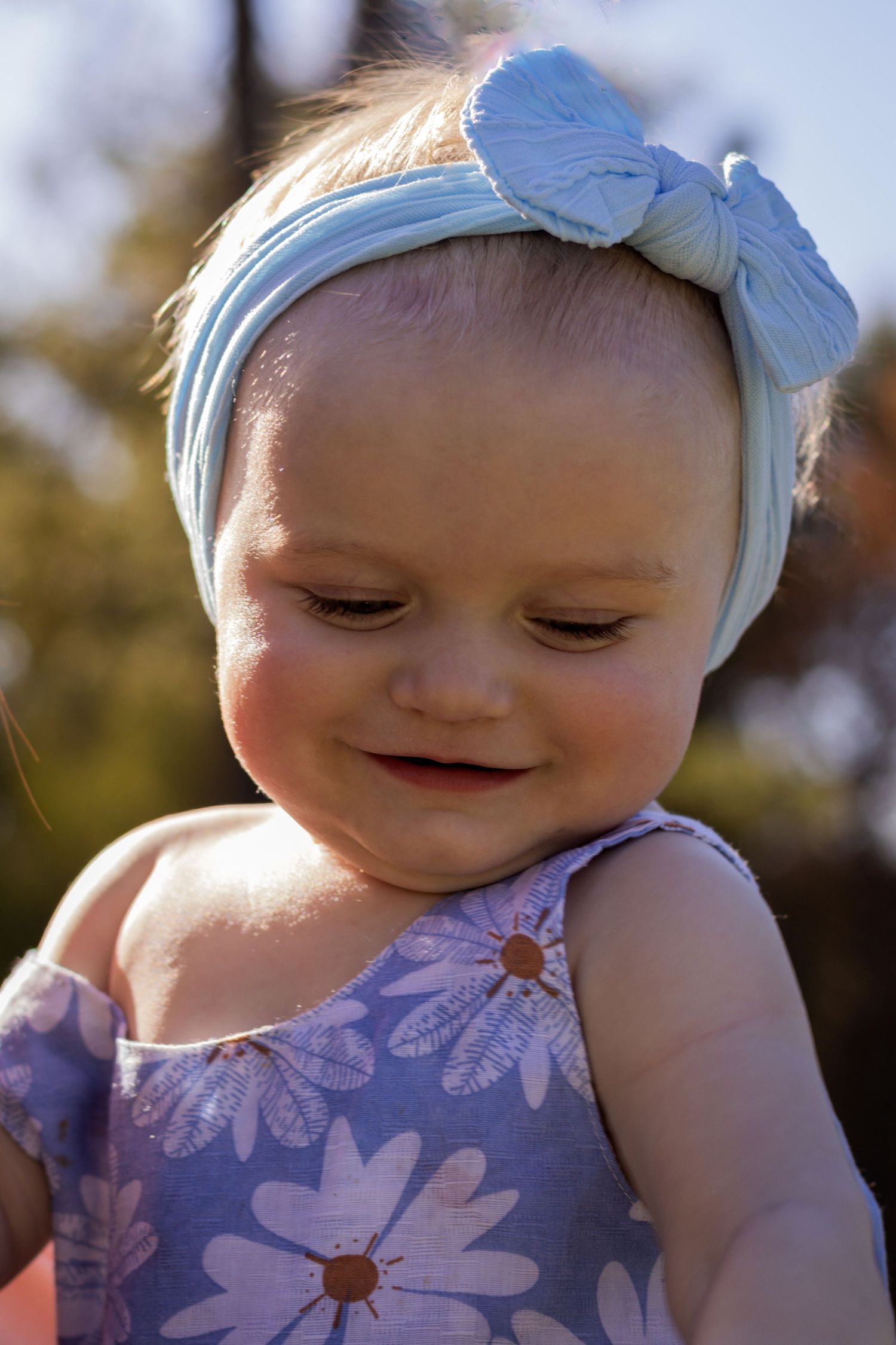 Baby wearing blue bow headband and floral dress smiles sweetly in summer sunlight.