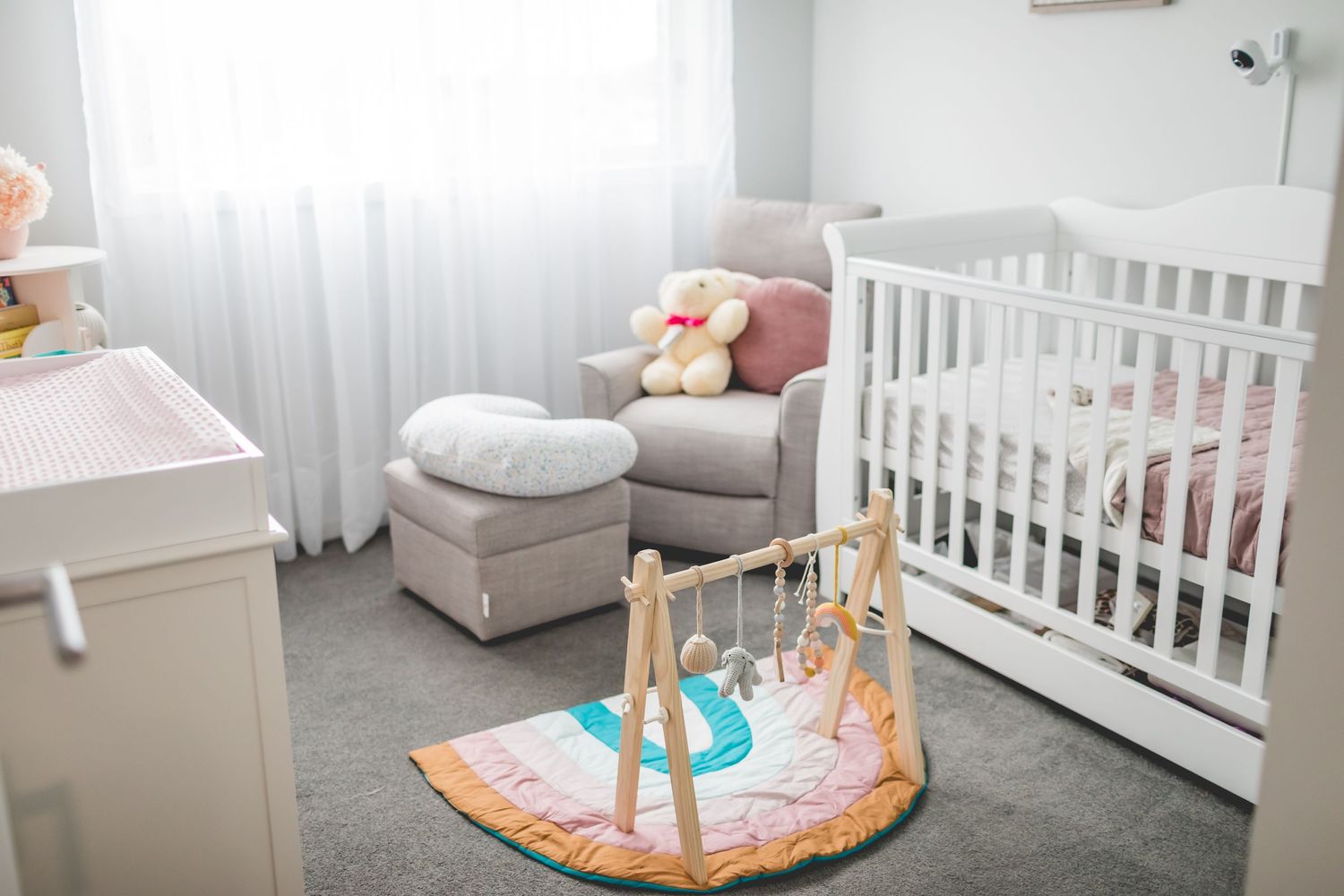 A cozy nursery with white crib, gray armchair, and colorful baby play mat.