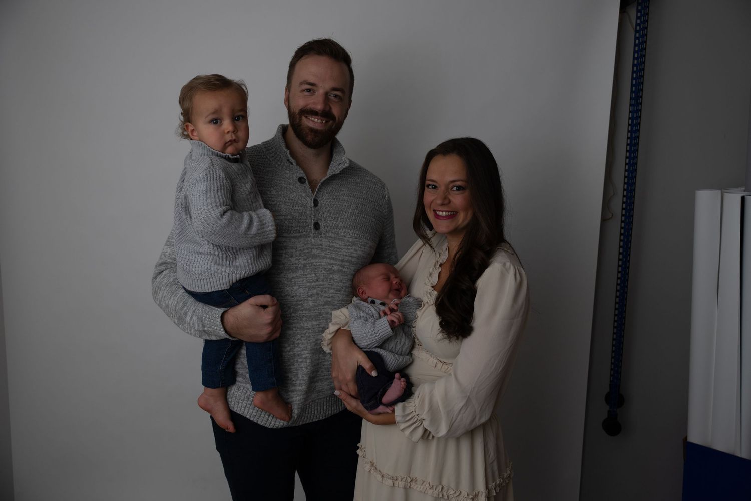 Indoor portrait of a young family with small children against neutral grey wall.
