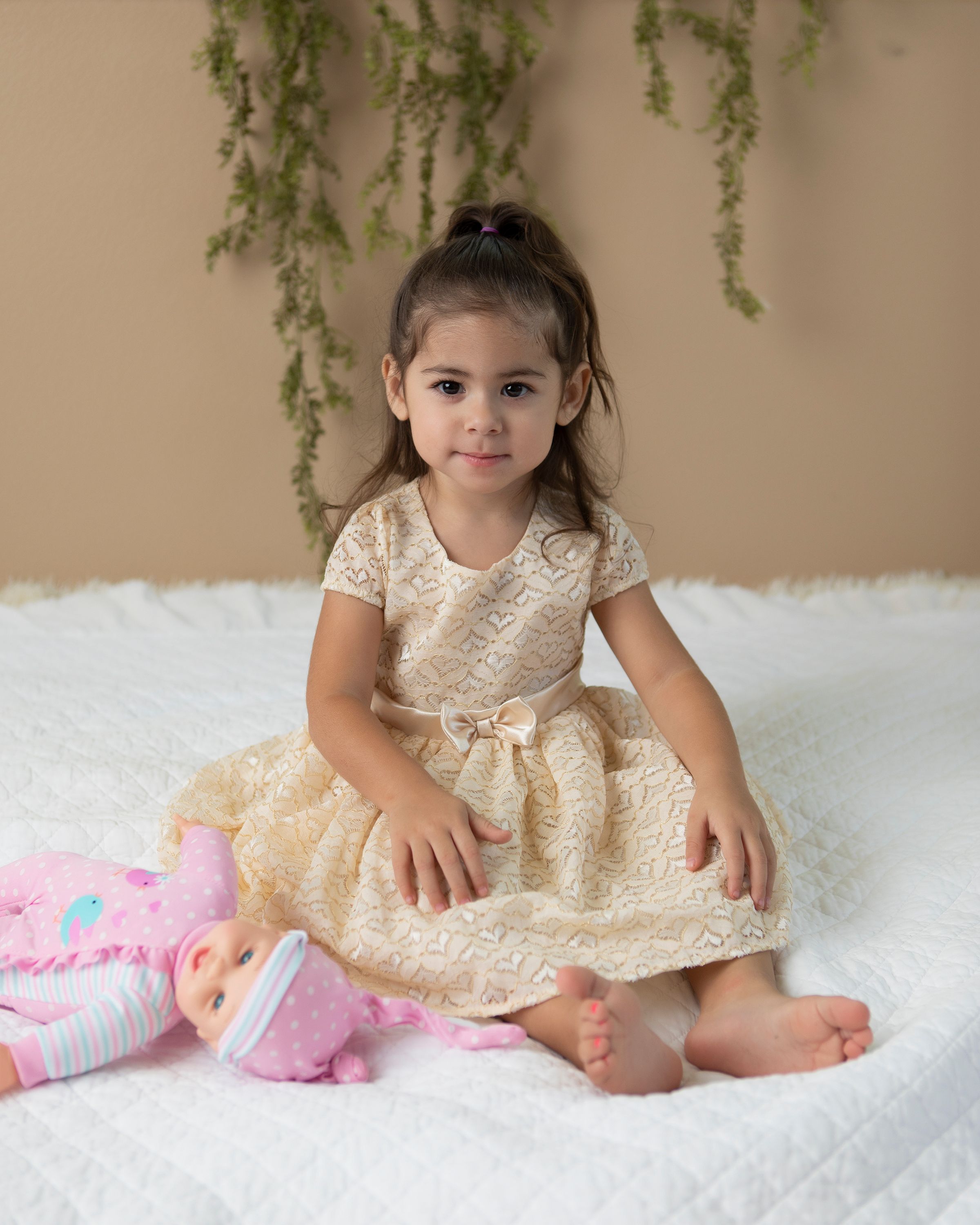 Child in gold lace dress sitting on white rug with pink toy during portrait session.