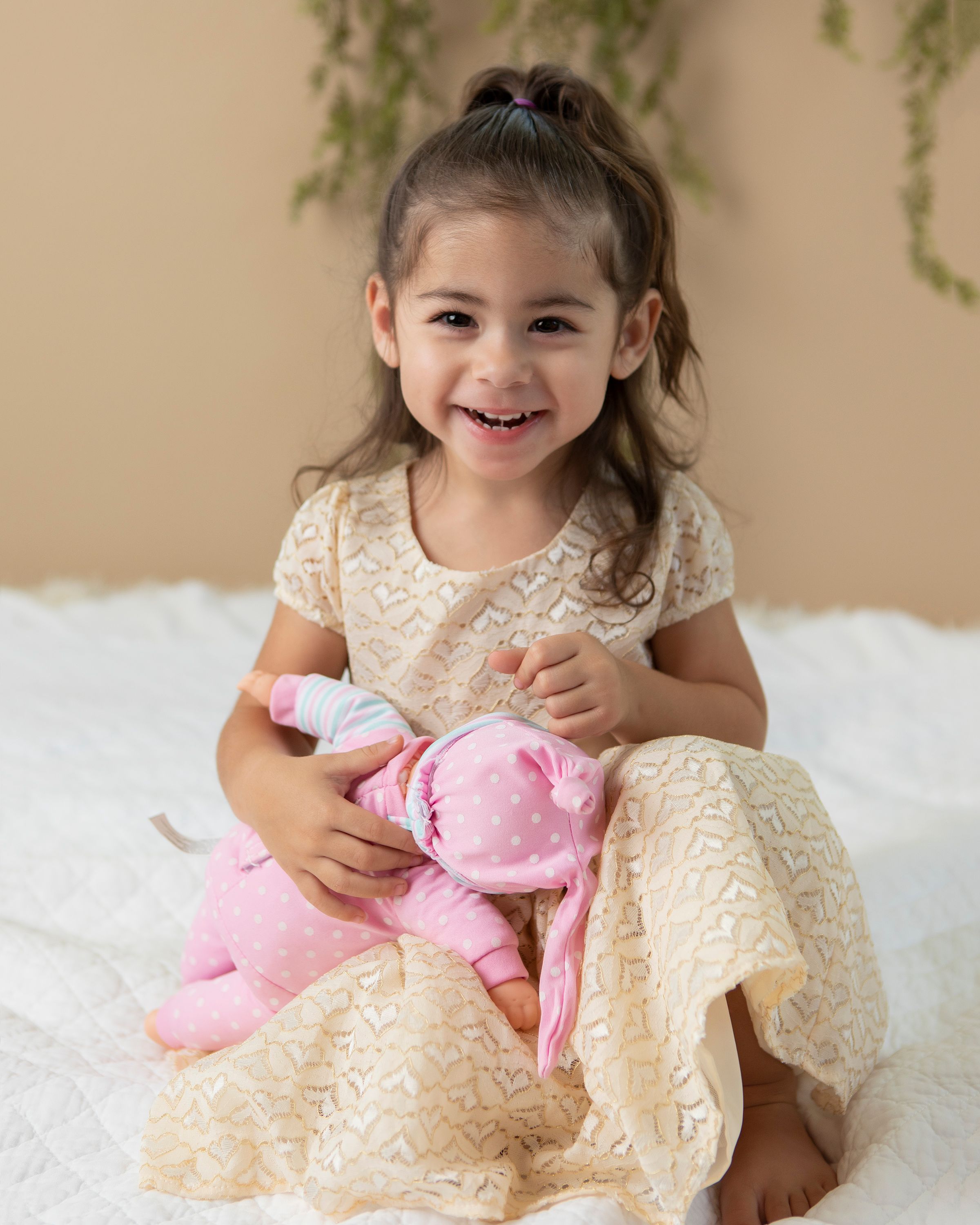 A small child in a cream lace dress smiles while holding a pink plush toy on a white fluffy rug.