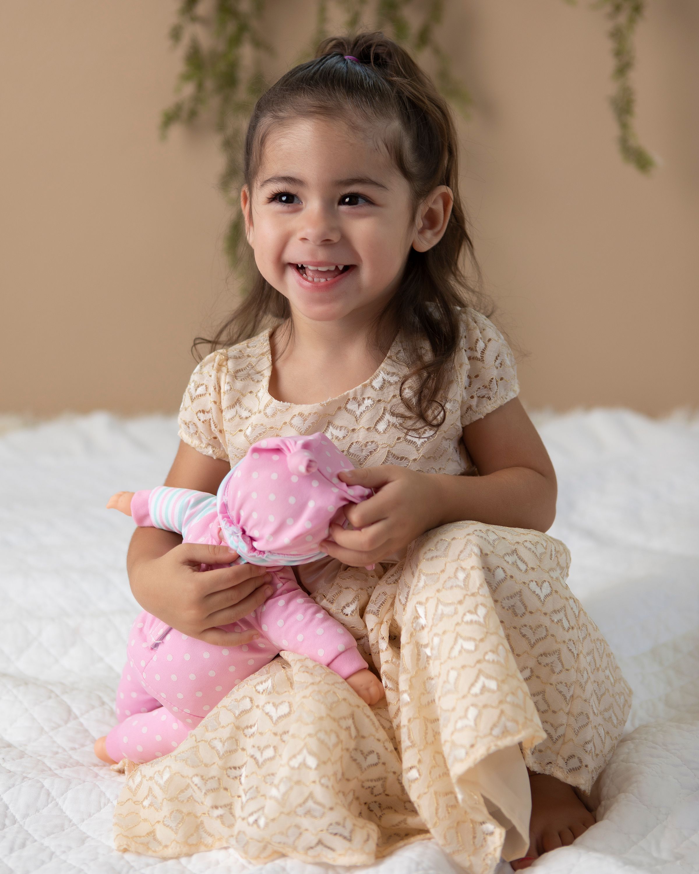 A child wearing a beige lace dress sits on white fur while holding a pink stuffed animal.