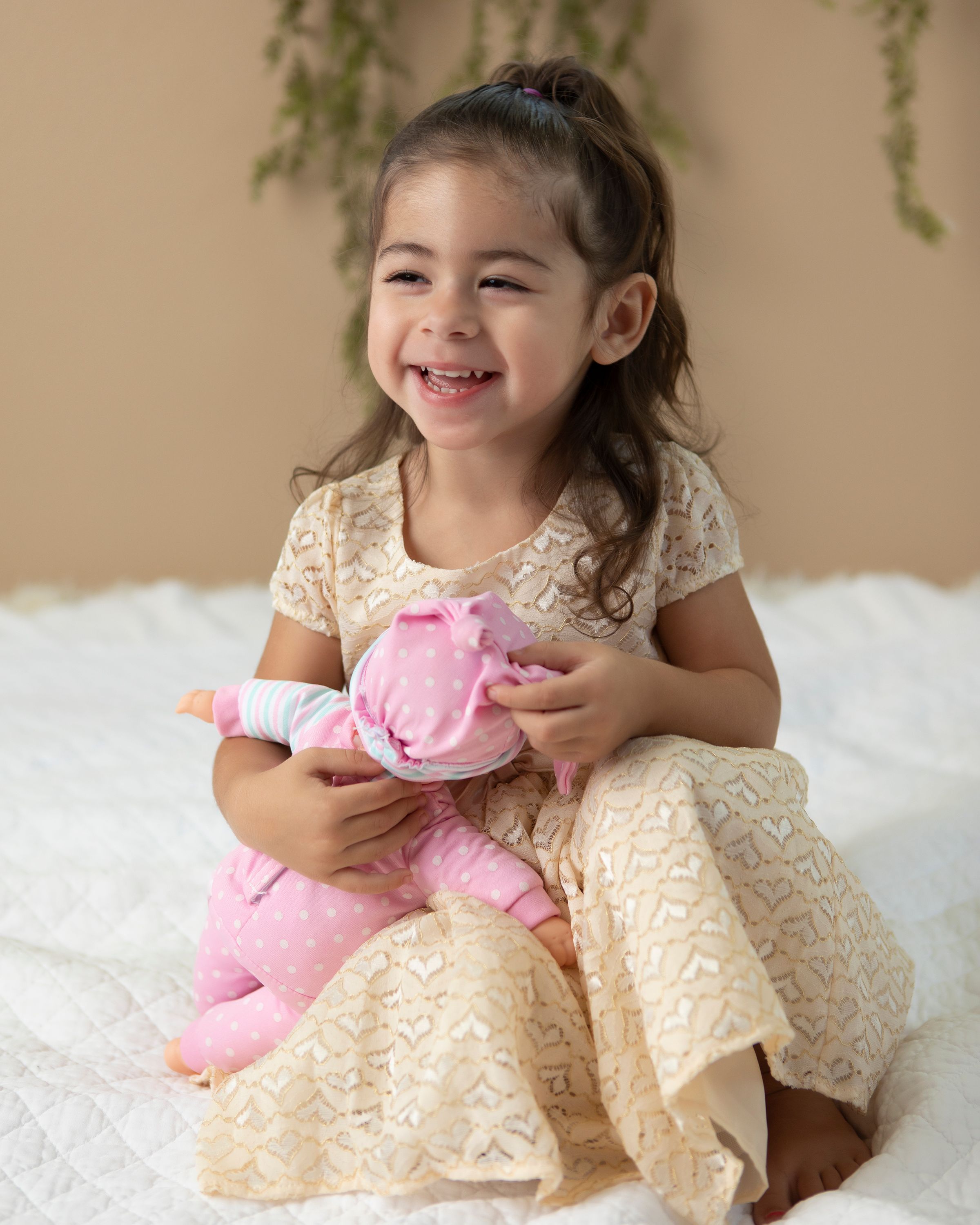 A happy child in cream colored dress holds a pink plush toy while sitting on white fur.