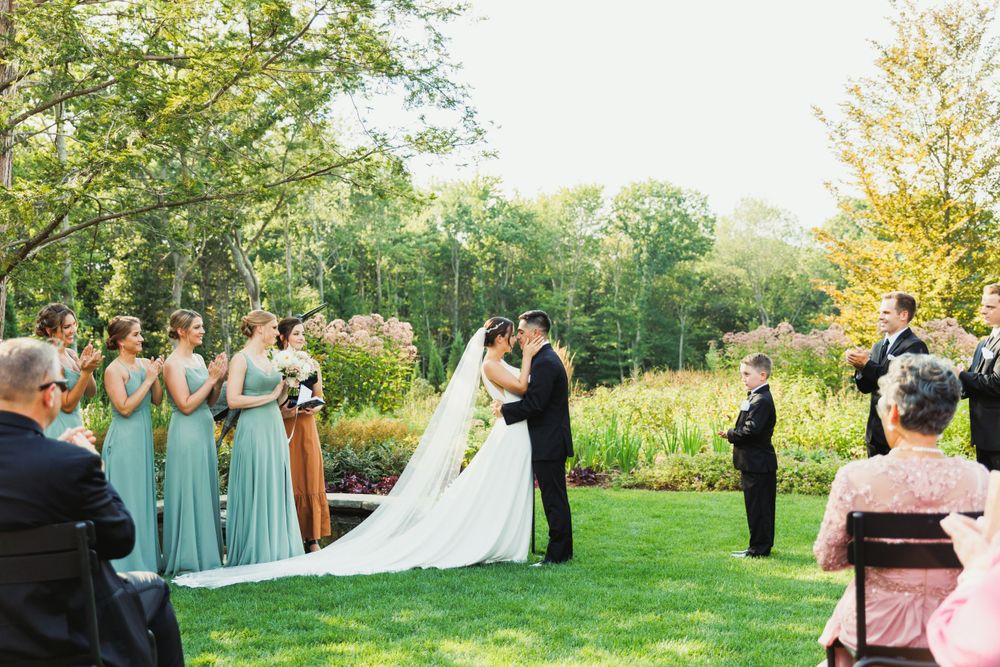 Wedding party standing on lush green lawn with bridesmaids in sage dresses during outdoor summer celebration.
