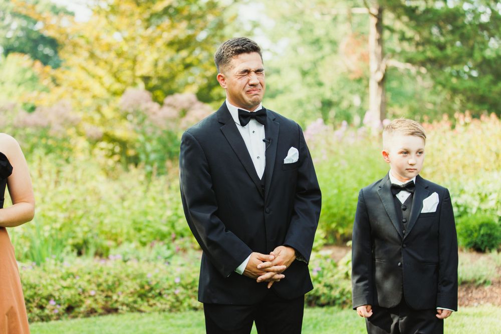Groomsmen in black tuxedos standing together during outdoor wedding ceremony.
