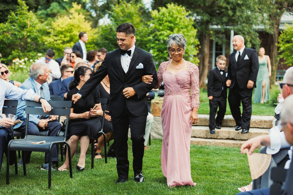 Wedding ceremony guests seated in white chairs on a manicured lawn surrounded by trees.