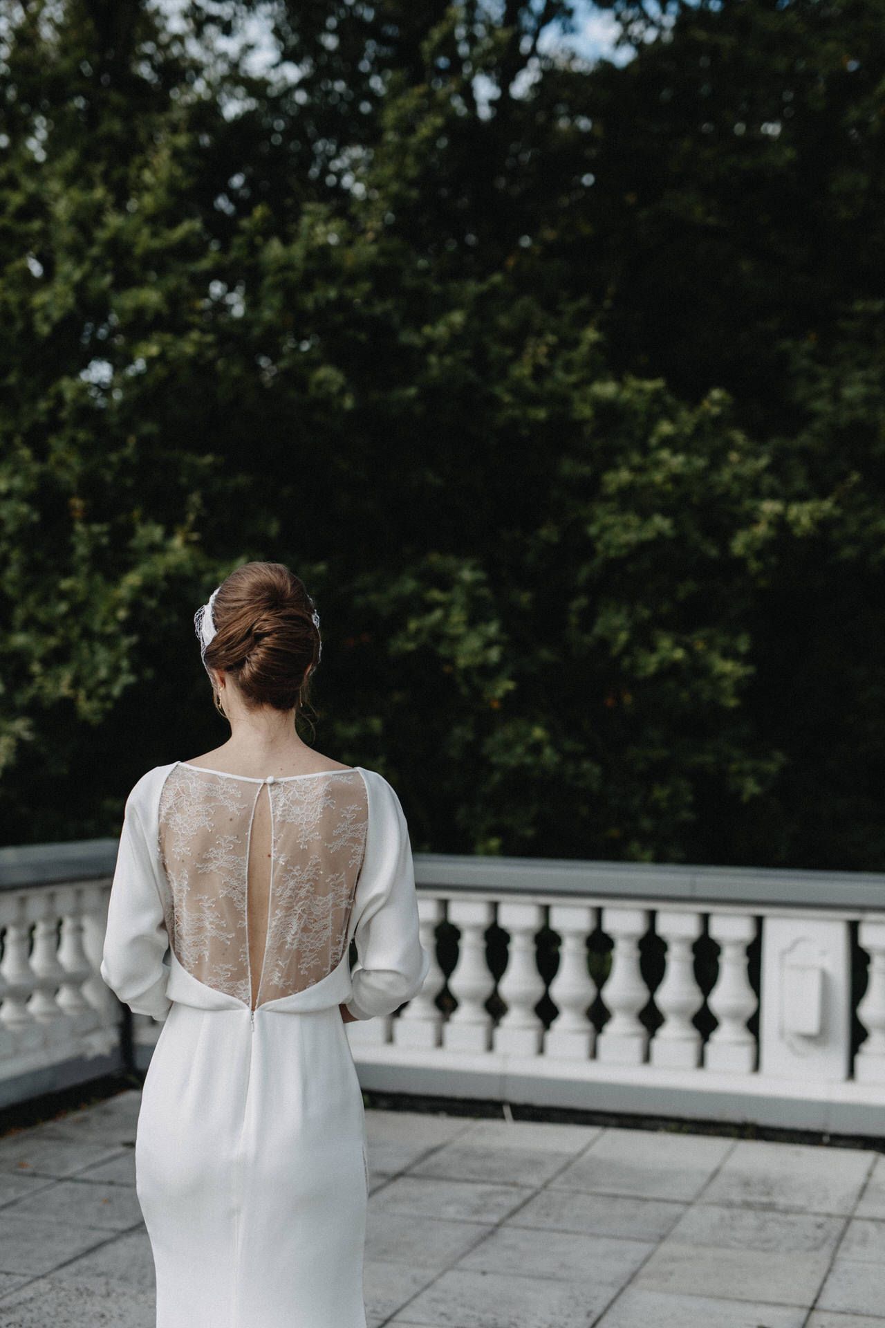 Back view of bride in elegant white dress with sheer lace panel standing on stone balcony.