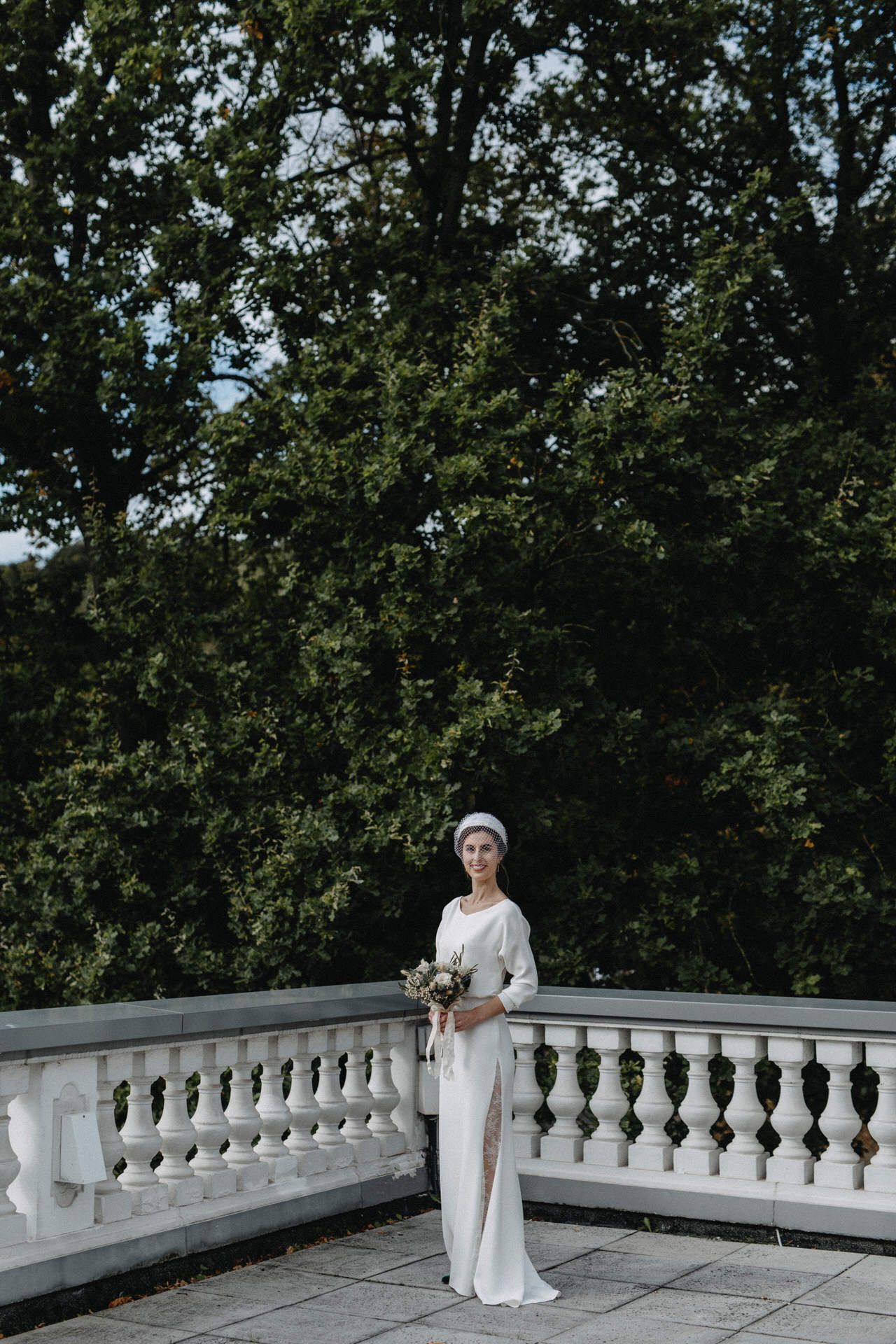 Elegant bride in white dress poses on classic stone balcony surrounded by lush greenery.