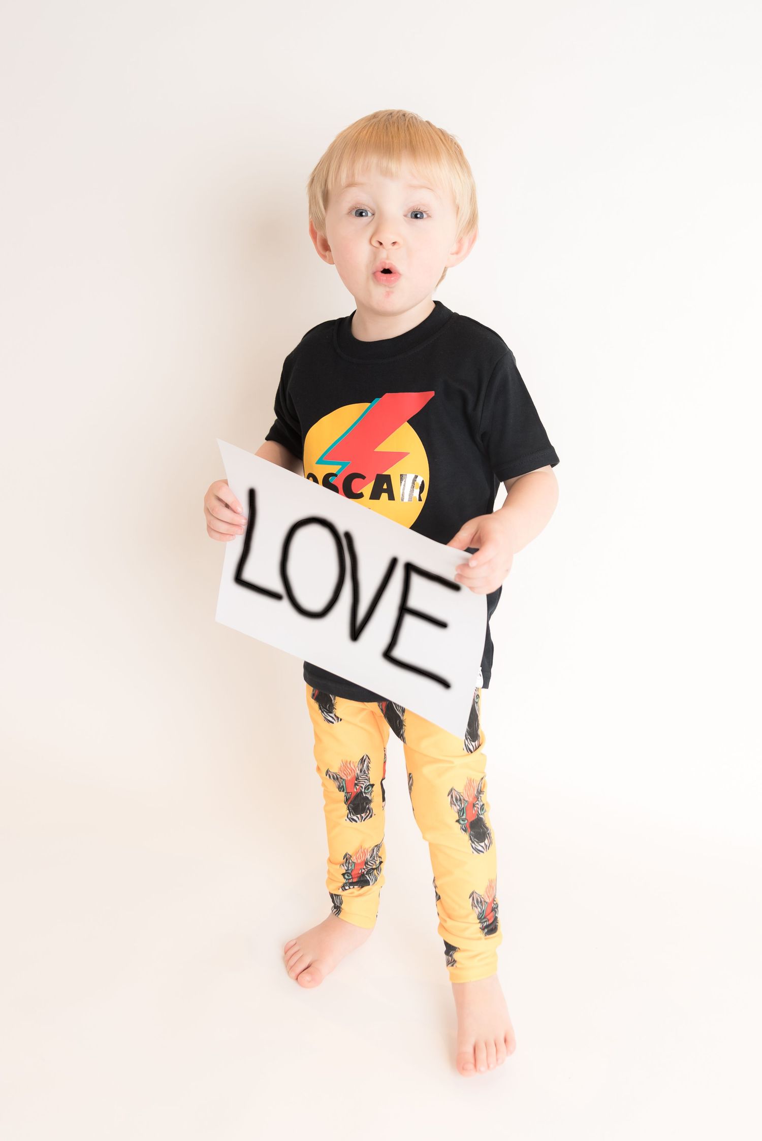 Young person in black t-shirt and yellow patterned pants holding a 'LOVE' sign.
