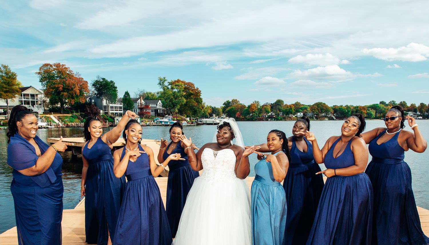 Bride and bridesmaids in navy blue gowns pose playfully on a dock with a scenic lake and fall foliage behind them.