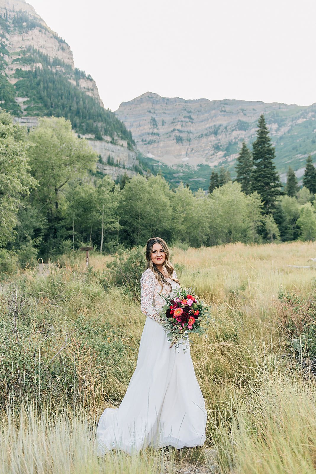 A wedding dress photoshoot in a mountain meadow with tall grasses and dramatic cliffs in the background.