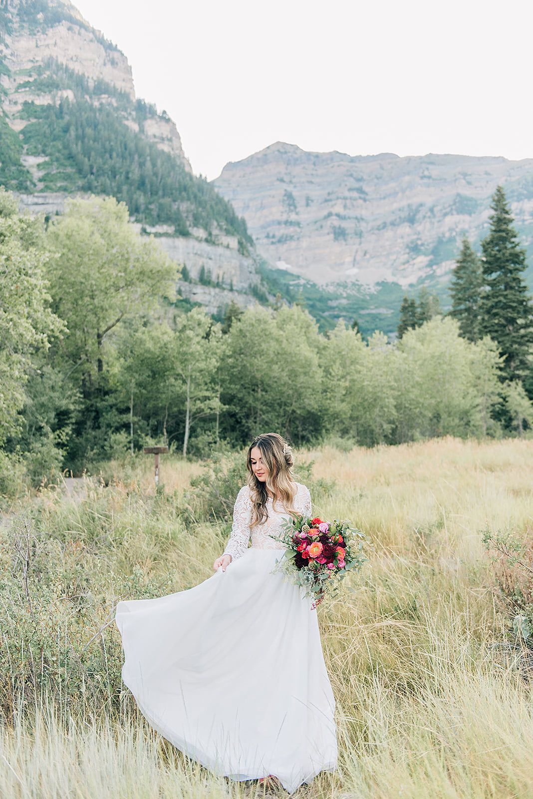 Wedding dress twirling in mountain meadow with dramatic mountain peaks and evergreen trees in background.
