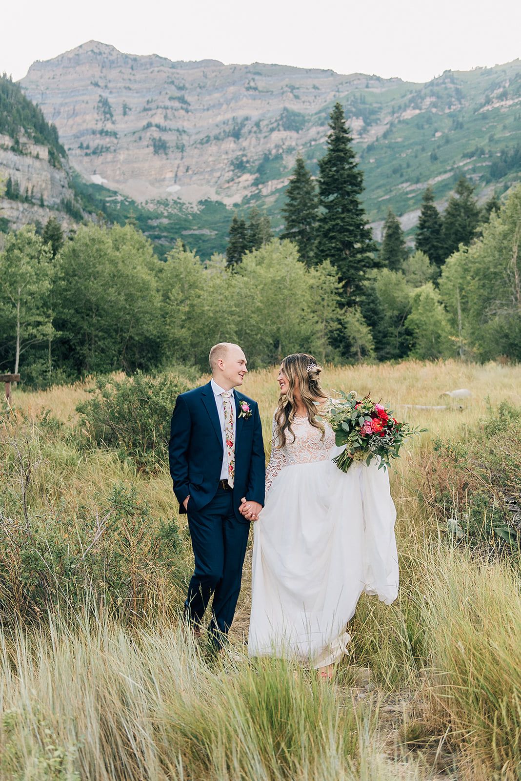 Wedding couple standing together in mountain meadow with colorful bouquet and dramatic mountain backdrop.