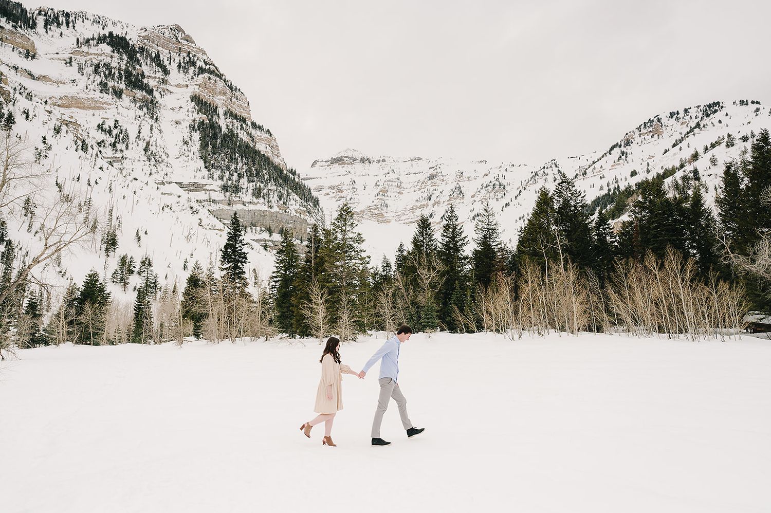 Couple walking hand in hand across snow-covered valley with mountain backdrop.