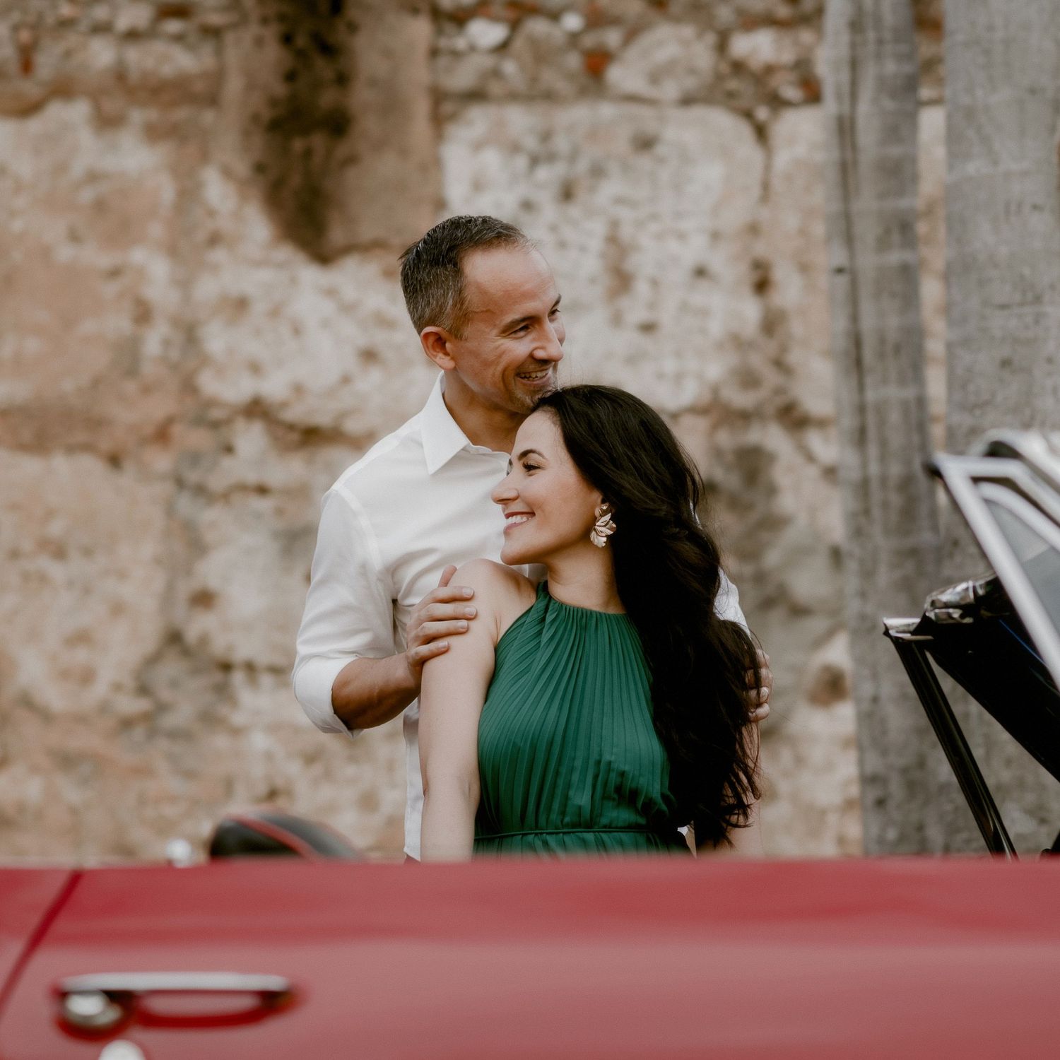 Romantic couple posing near vintage red car against stone wall.