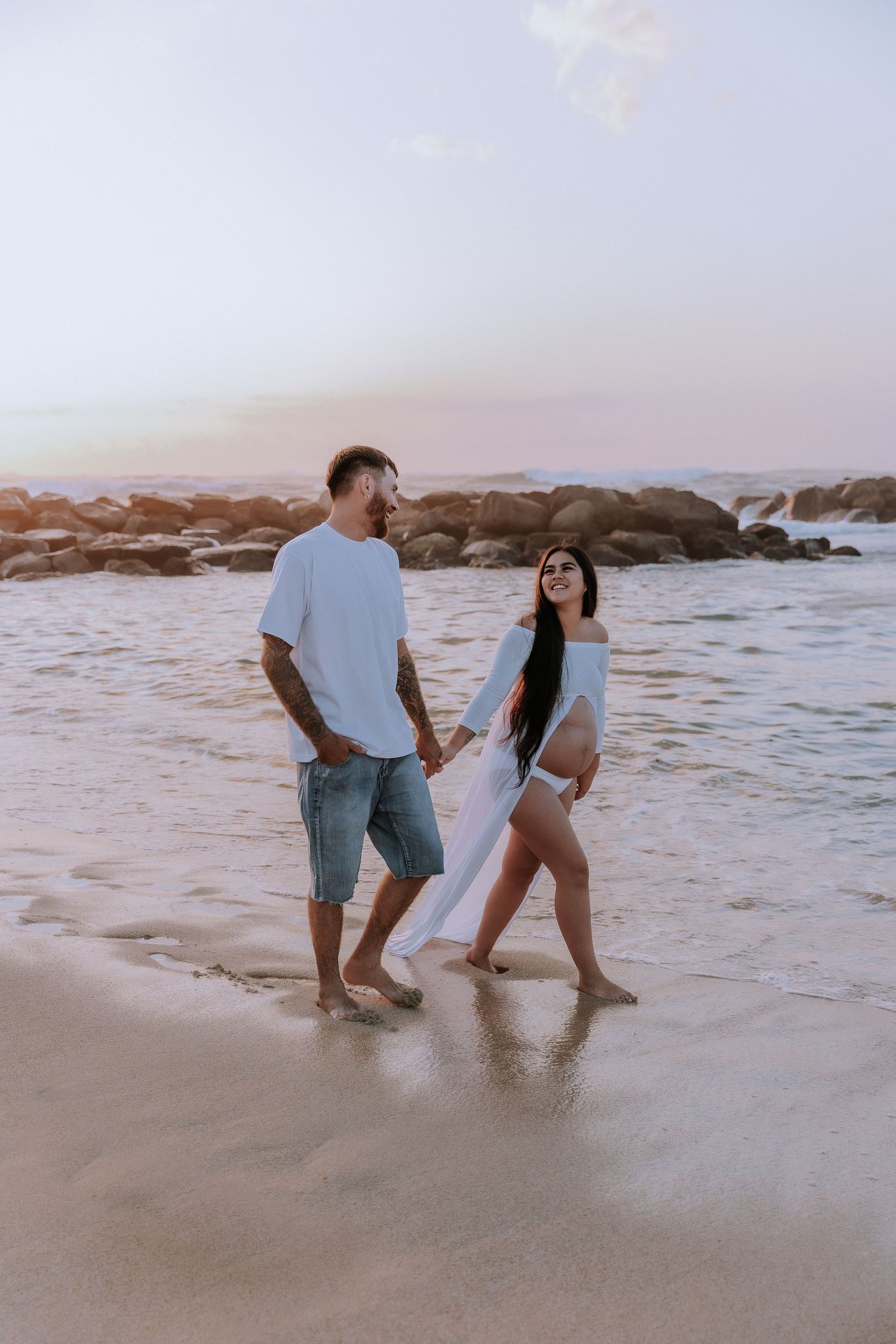 A couple walking together on a beach at sunset with waves crashing nearby.
