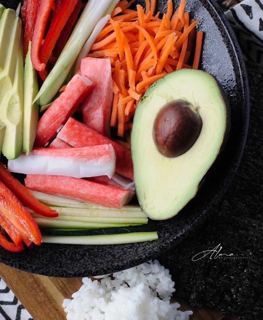 Fresh sushi ingredients including sliced avocado, carrots, watermelon and vegetables arranged on a black plate with rice.