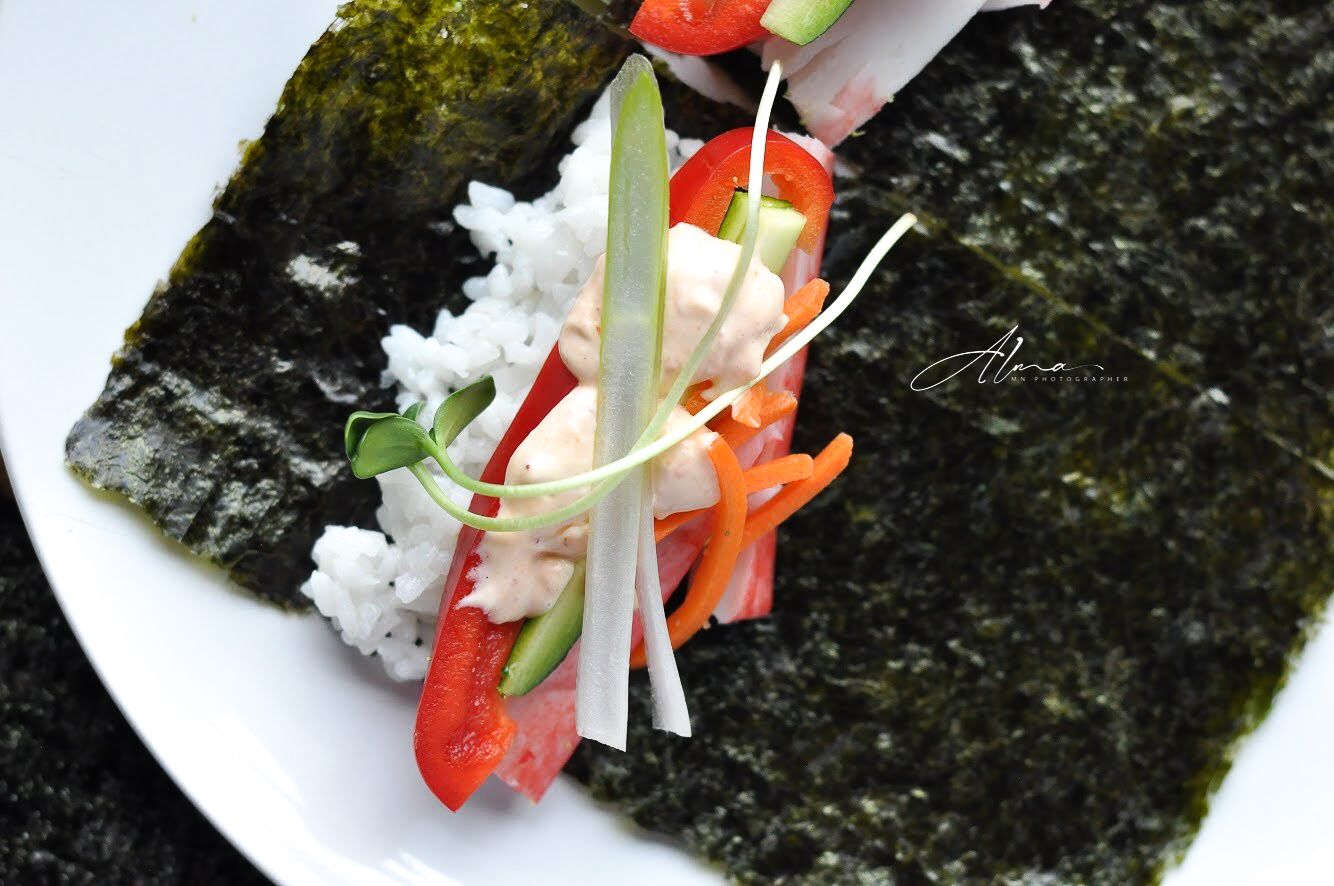 Close-up of sushi hand roll with rice, vegetables and nori wrapper on white plate.