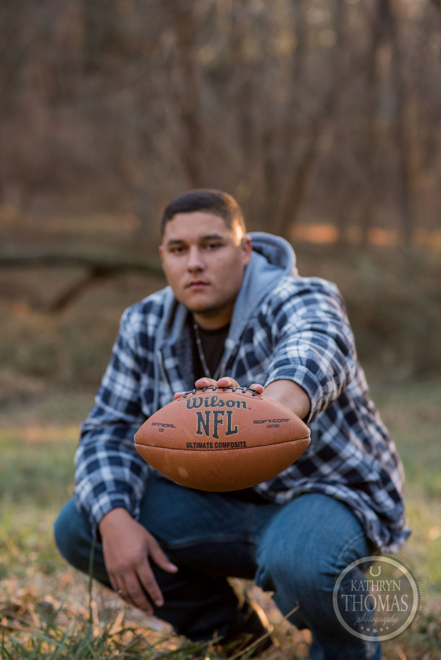 Someone in plaid holds an NFL football while crouching in a wooded outdoor setting at sunset.