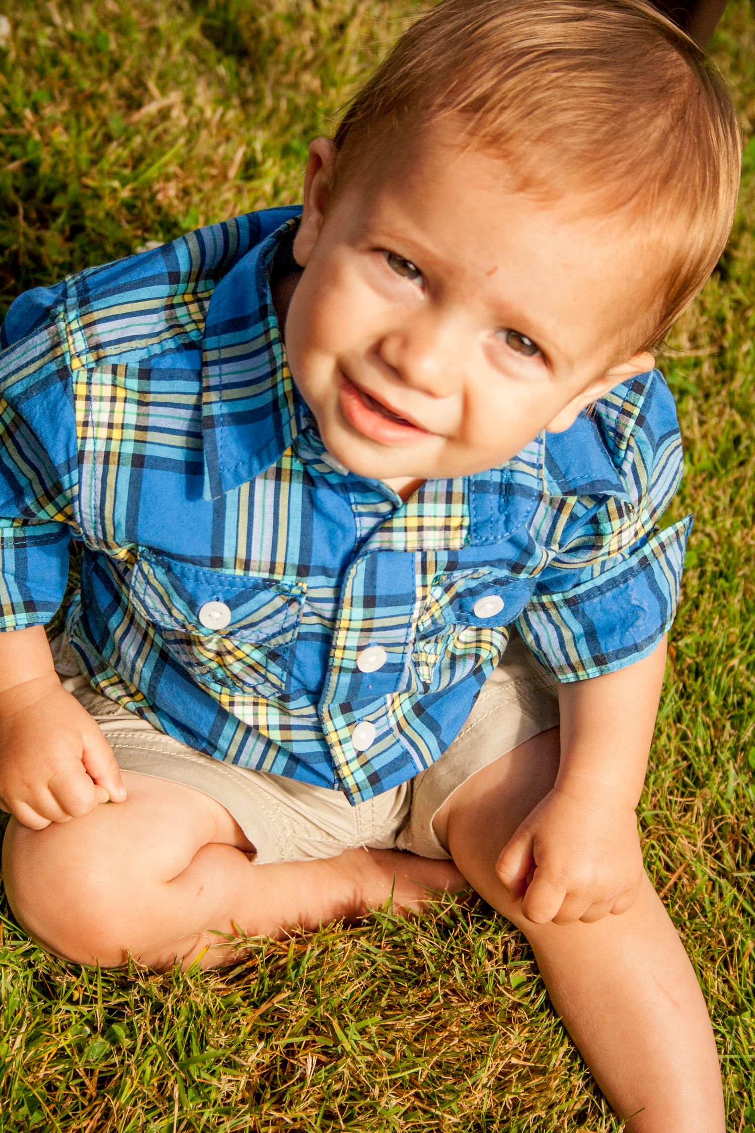 Young child in blue plaid shirt sitting on grass outdoors.