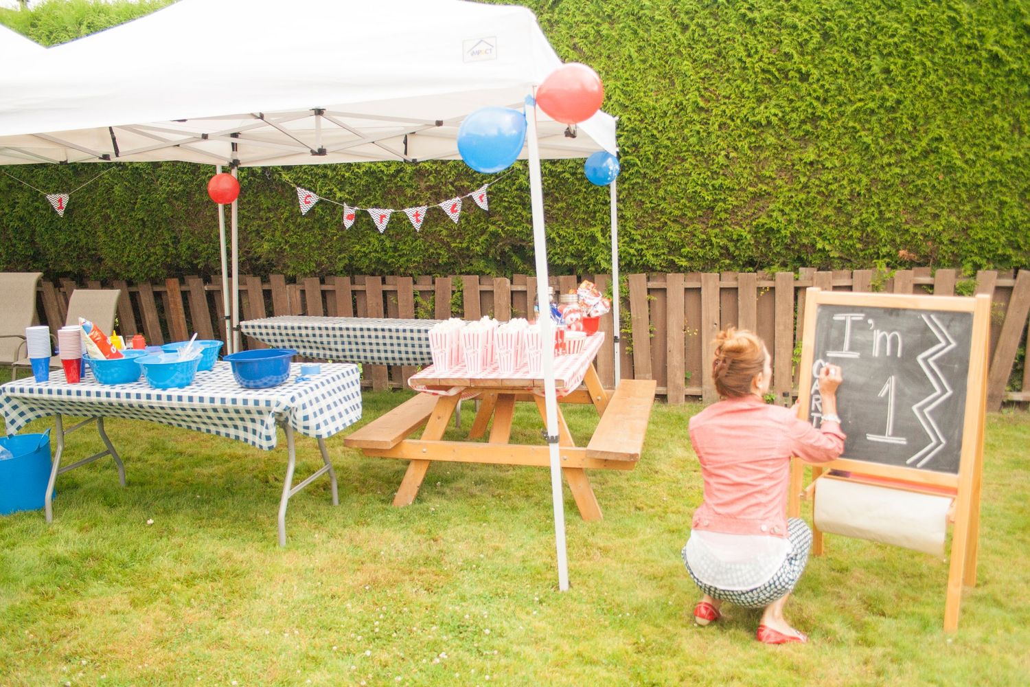 Outdoor party setup with picnic table, balloons, and chalkboard in a backyard garden.