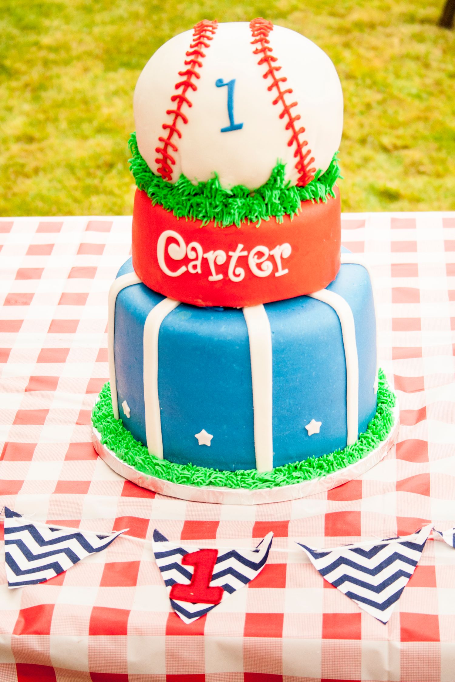 Baseball themed birthday cake for Carter with red and blue tiers on checkered tablecloth.