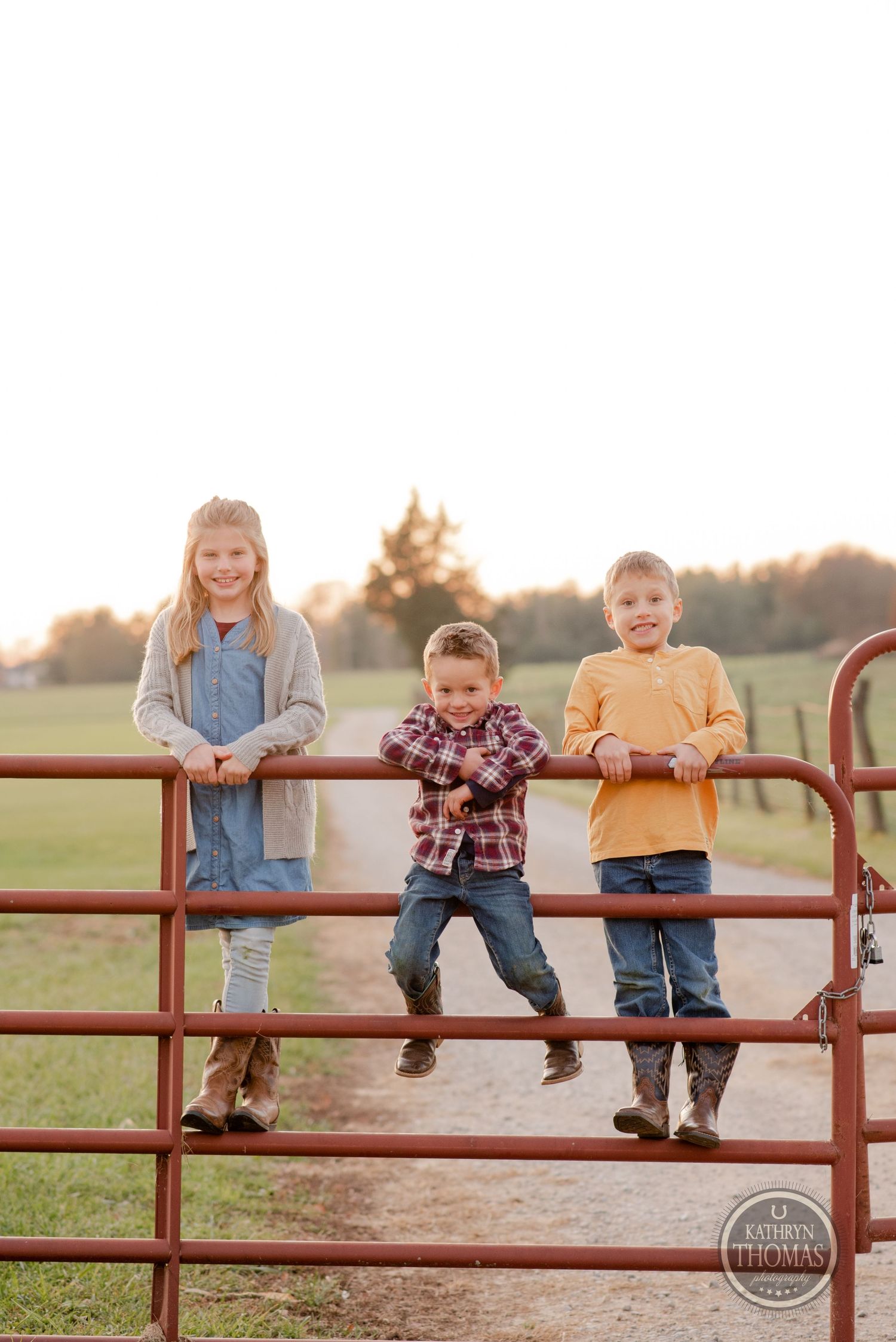Three children perched on a red metal farm gate at sunset in a rural setting.