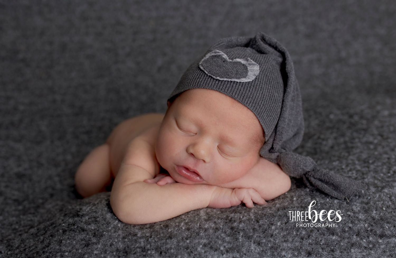 Sleeping newborn wearing a grey knit hat with heart detail on dark textured background.