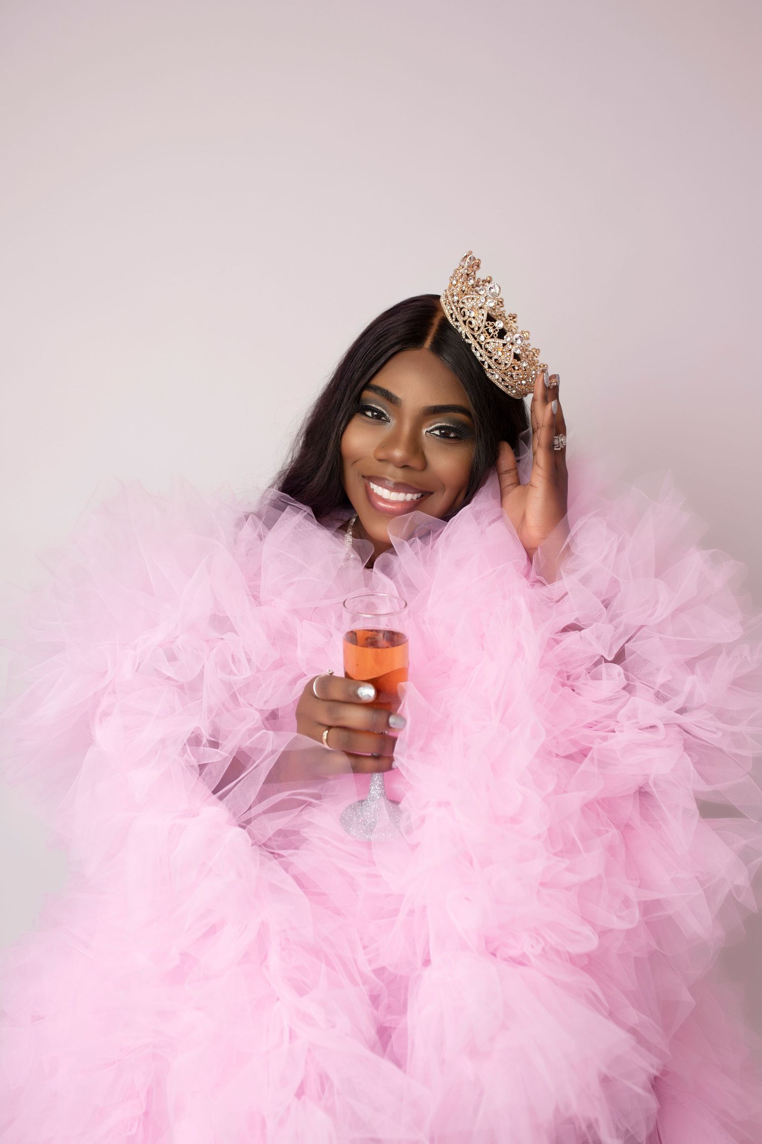 Glamorous portrait in pink tulle gown with crown and champagne glass against light background.