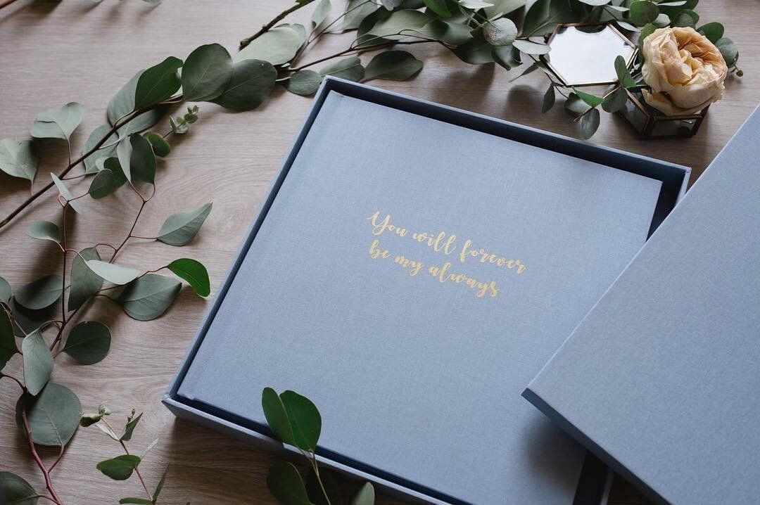 Gray photo album with gold lettering surrounded by eucalyptus branches and flowers.