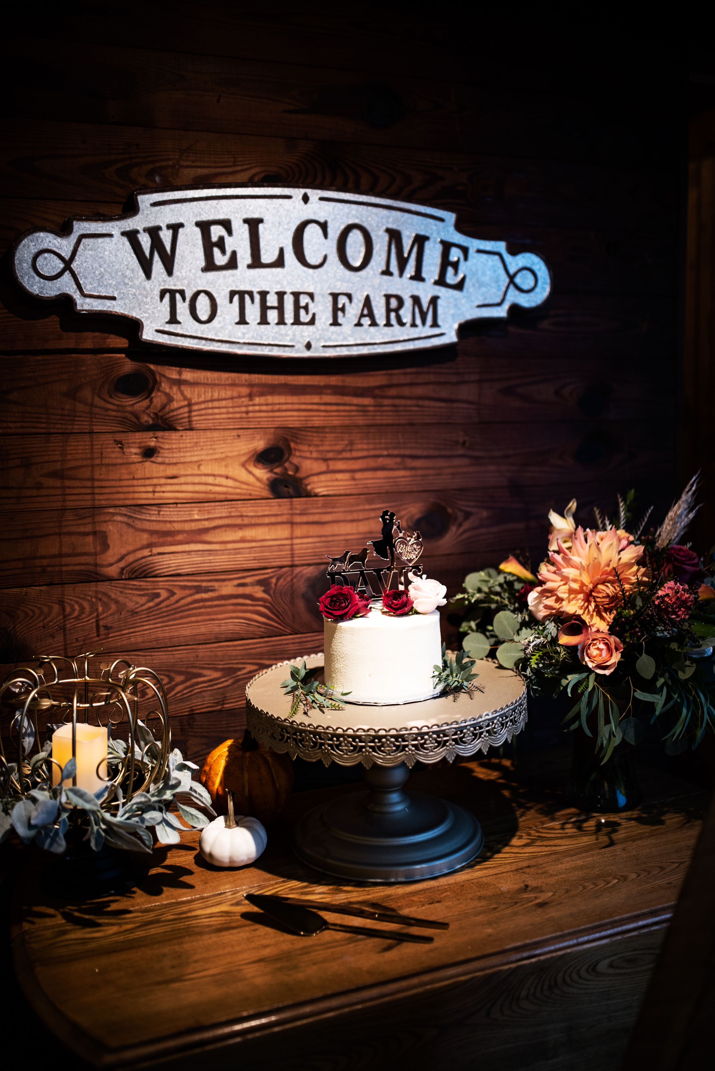 Rustic farm wedding cake display with welcome sign and floral arrangements on wooden table.