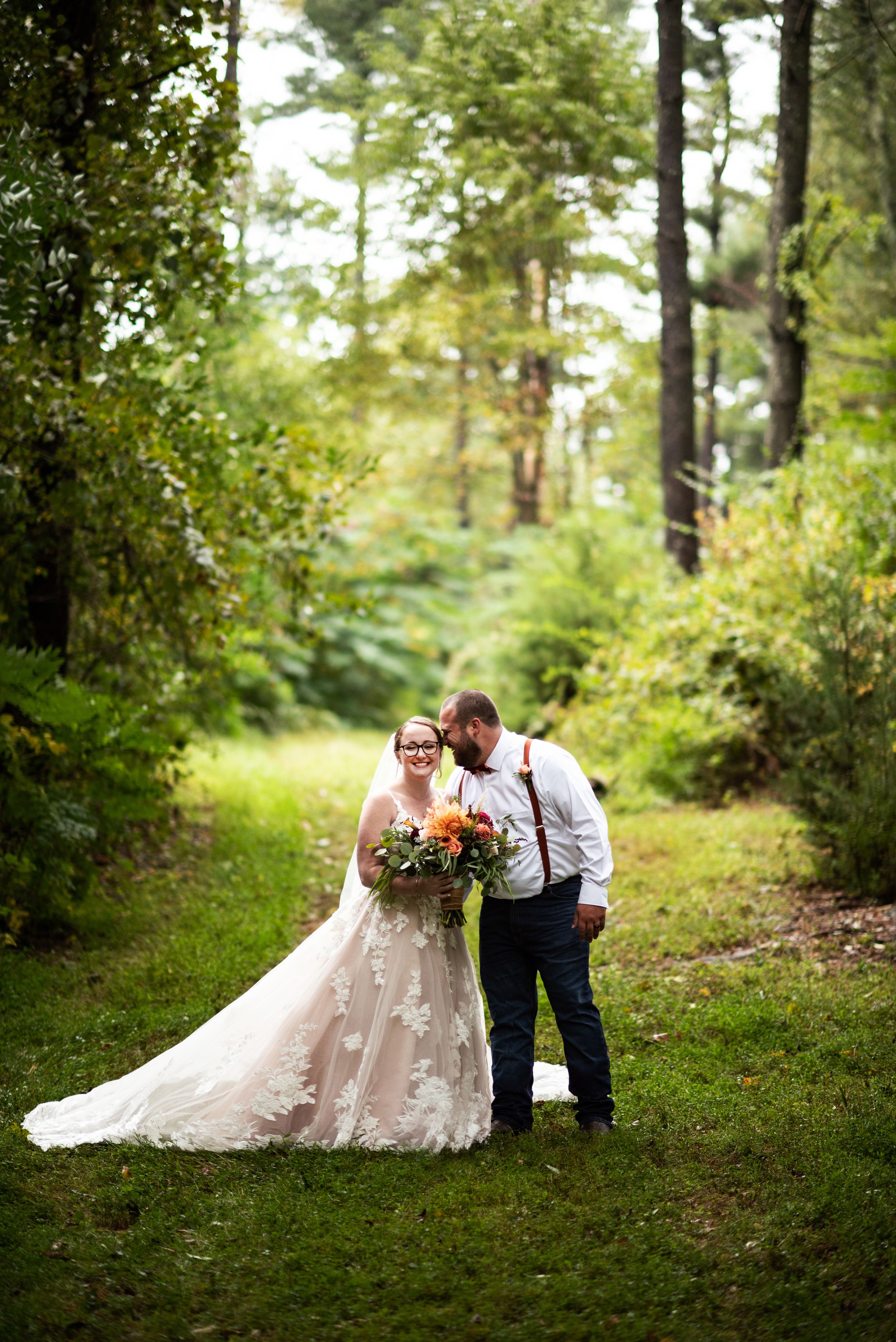 A romantic wedding couple shares an intimate moment on a tree-lined forest path.