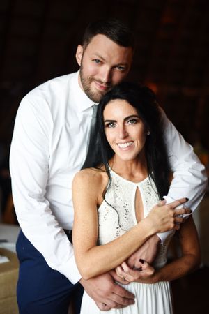 Happy couple embrace in formal attire with woman wearing white lace dress while posing indoors.