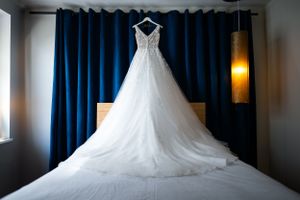 A beautiful white wedding dress with detailed lace hanging against blue curtains in natural light.