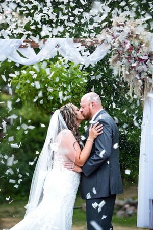 Wedding ceremony kiss under white arch with confetti falling.