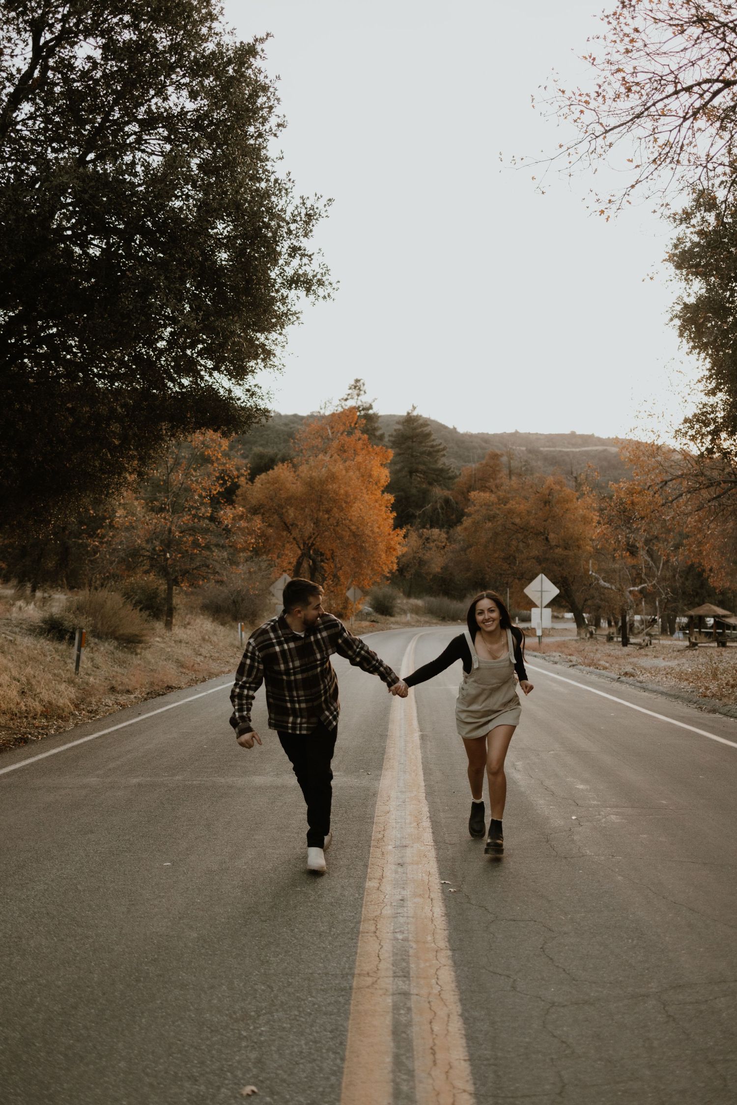 Two people hold hands while walking down the middle of an empty road surrounded by autumn trees.