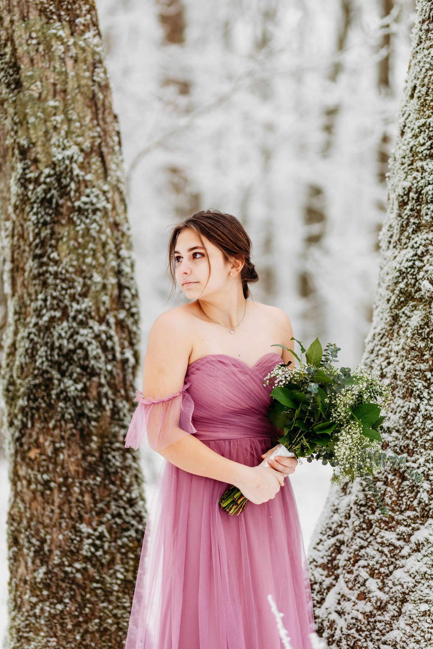 Woman in purple strapless gown holding bouquet between snow-covered trees.
