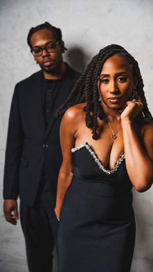 Stylish couple in formal black attire pose against gray wall for elegant portrait.