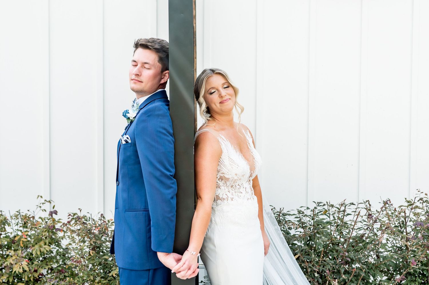 Wedding couple poses against white wall with green shrubs in elegant attire.