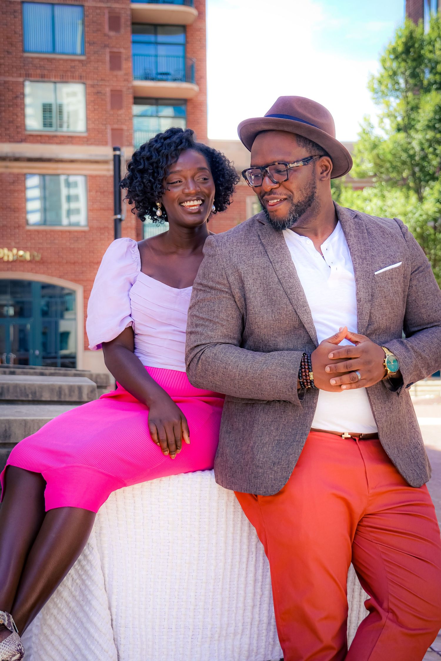 A stylish couple sitting together outdoors in vibrant pink and orange outfits.