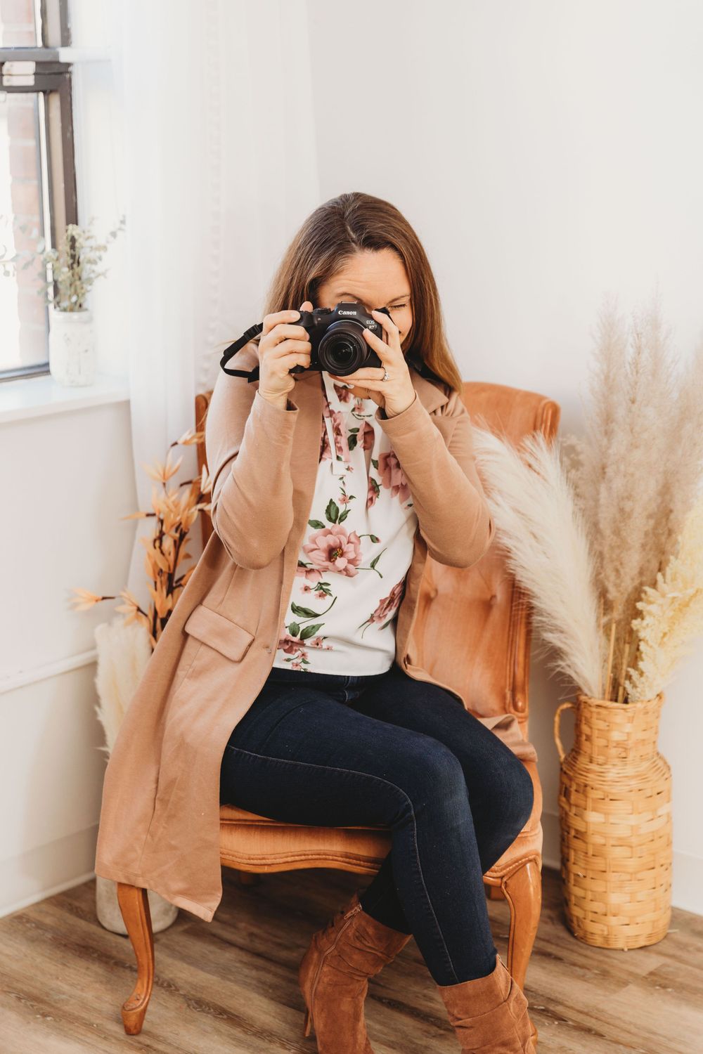 Professional photographer sitting in a tan leather chair with a camera and floral top against a bright interior.
