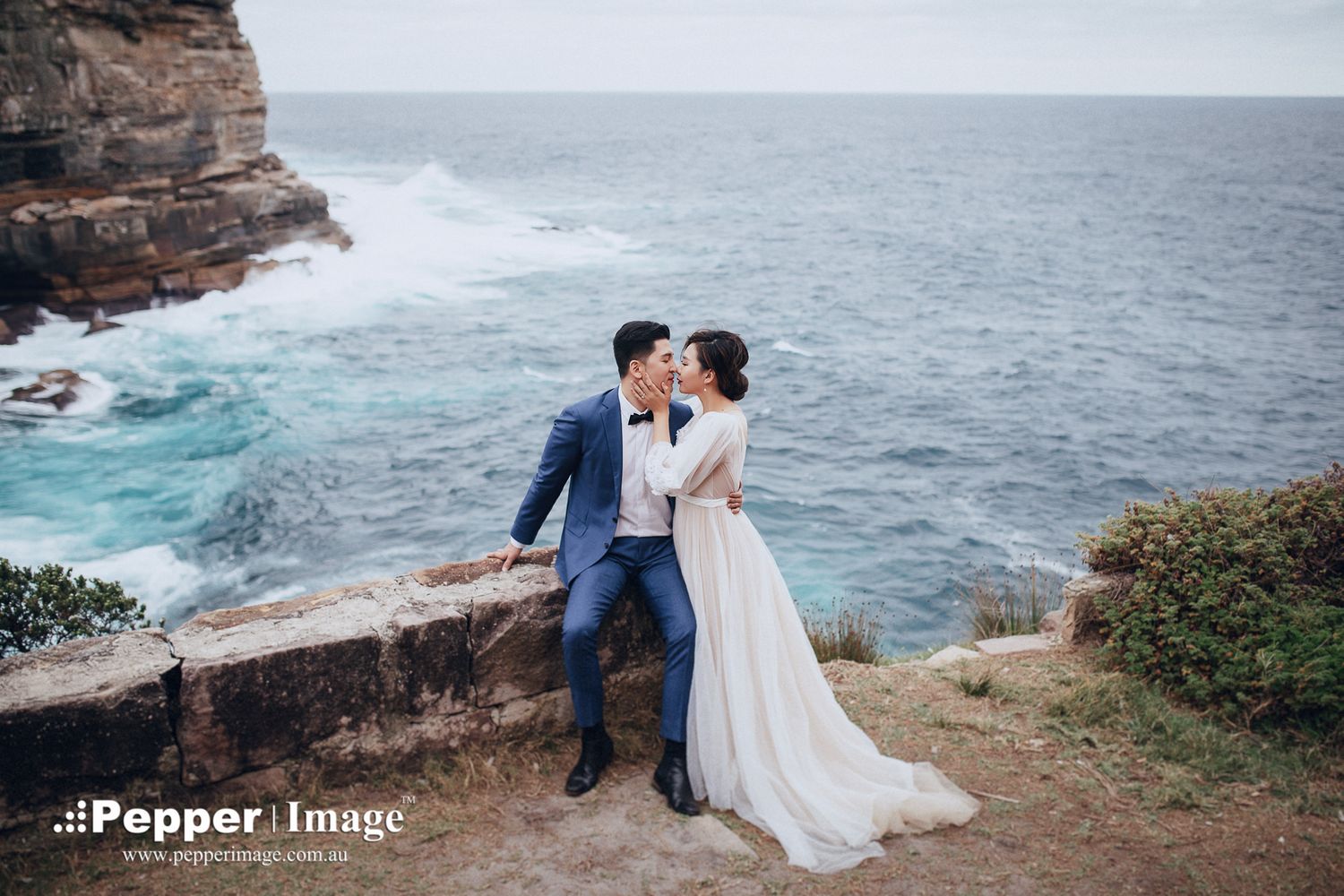 Couple sharing romantic kiss on stone wall overlooking dramatic ocean cliffs and waves.