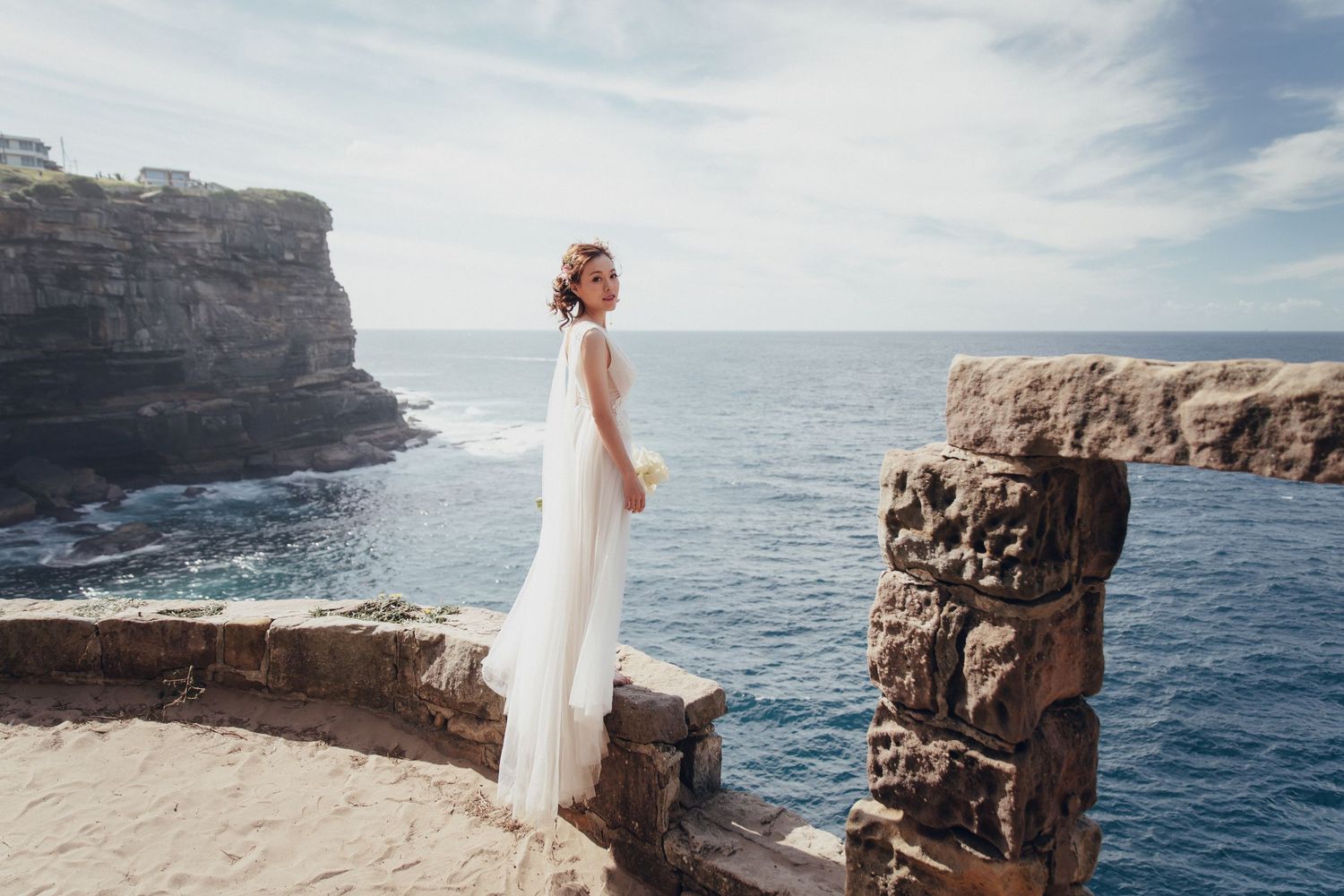 Bride in flowing white dress standing on rocky coastal cliff with ocean backdrop.