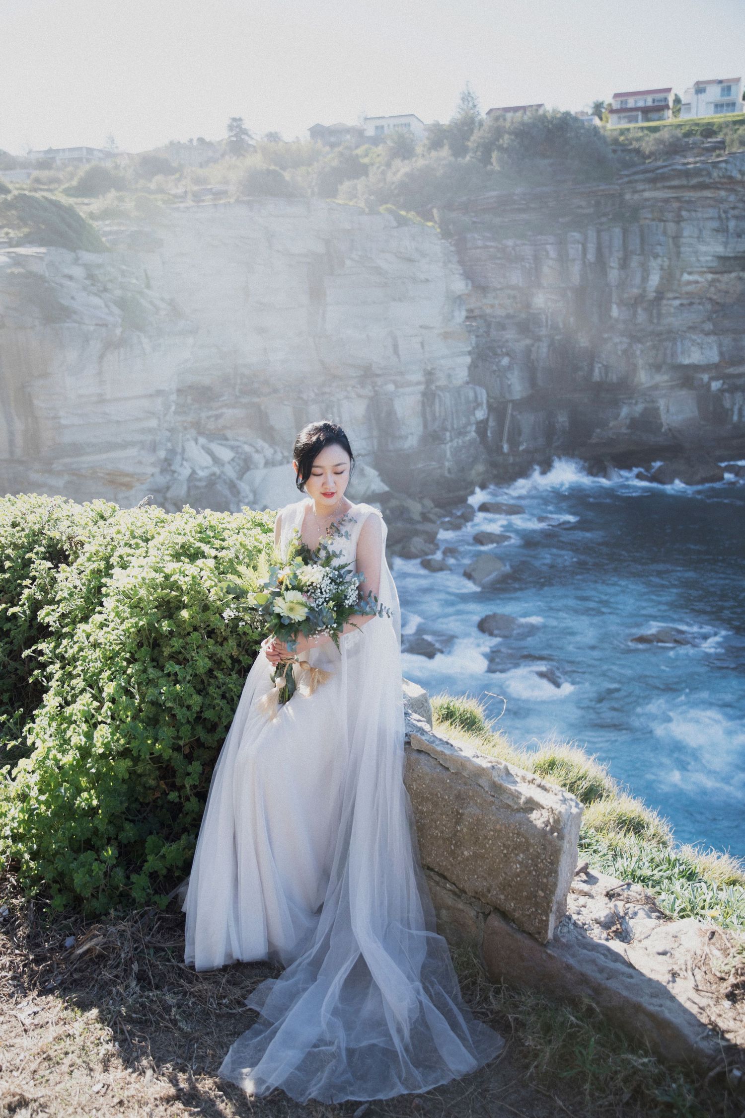 Bride in grey tulle gown holding white bouquet on coastal cliff with ocean view.