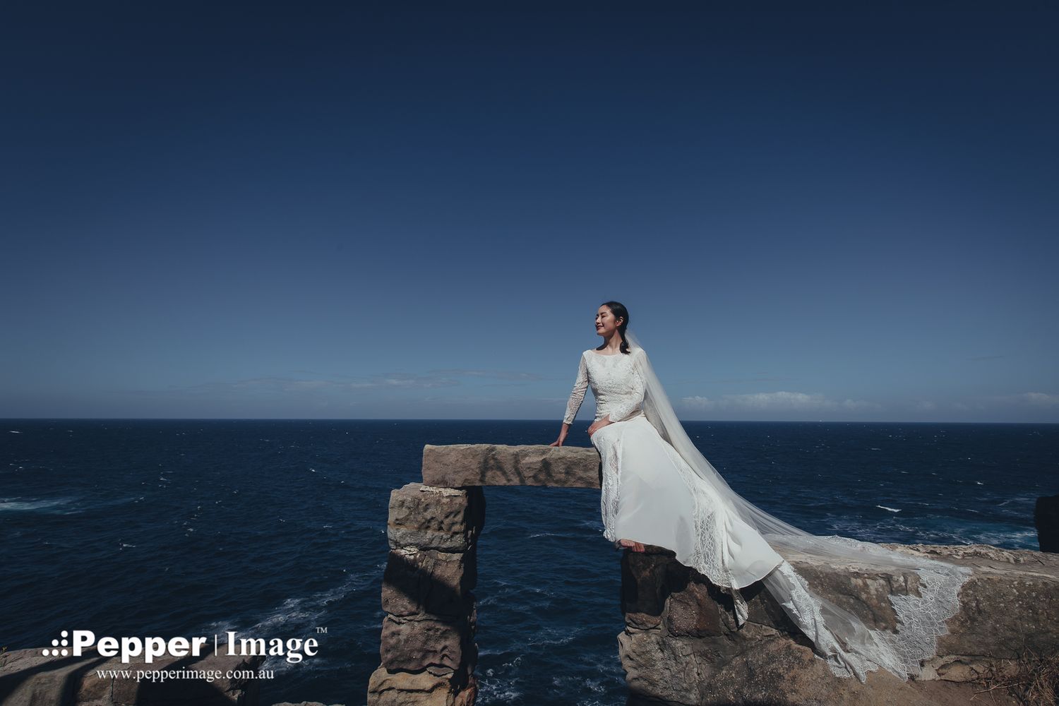 Bride in white gown with veil sitting on stone pier against deep blue ocean sky.